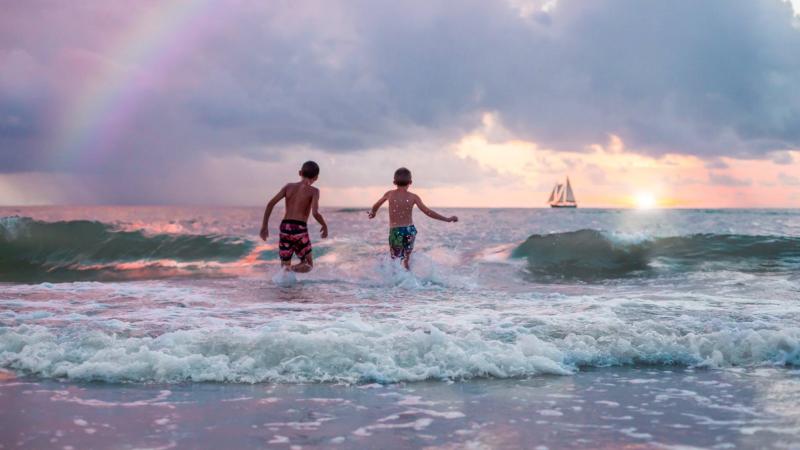 Two teenage boys running into the waves in the late afternoon, framed by a distant sailboat and part of a rainbow.