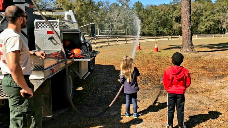 A young girl spraying water from a fire hose while a young boy and a Ranger watch. 