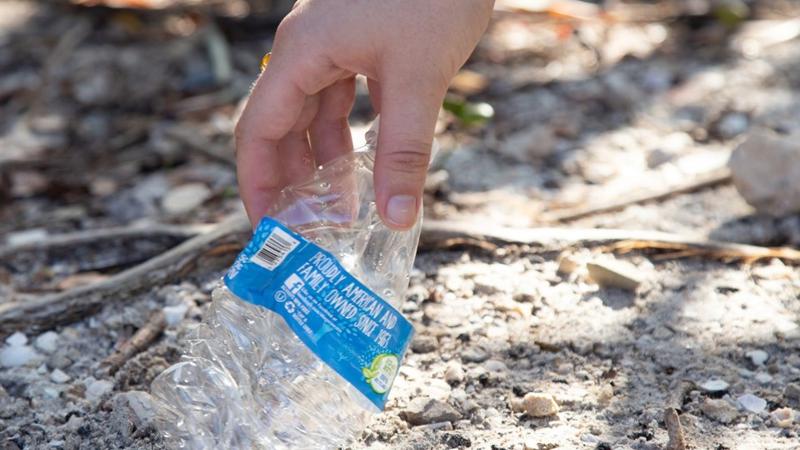 A hand reaches down to pick up a crumpled plastic water bottle. 