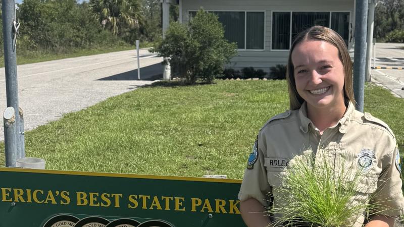 A smiling woman in park ranger uniform, holding a pot of long grass. 