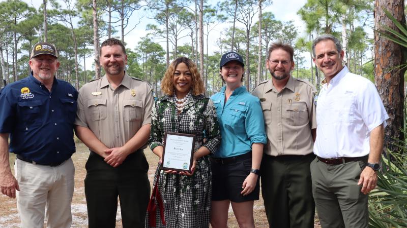 Six people standing shoulder to shoulder. A woman in the middle is holding a plaque