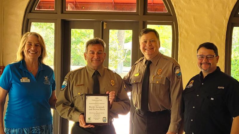 A group of four people standing shoulder to shoulder, a park ranger second from the left is holding a plaque