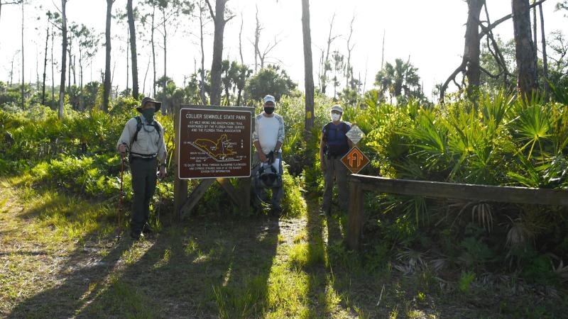 Three hikers standing by a hiking trail sign with green trees and grass.