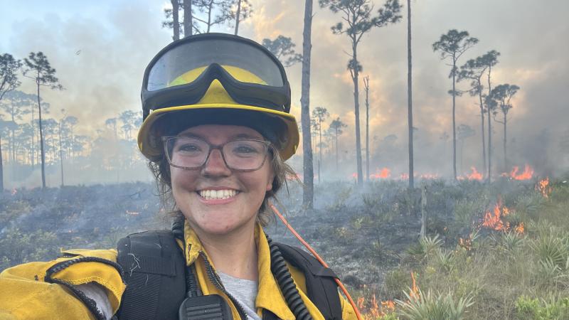 A smiling woman in fire PPE, standing in front of a prescribed fire. 