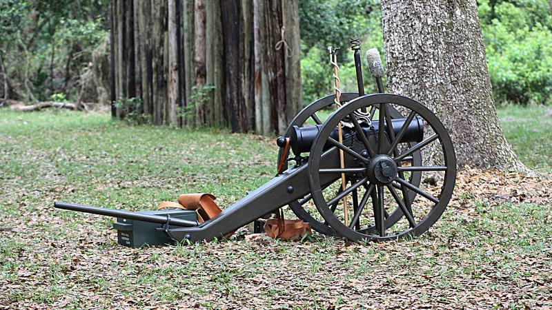 a small cannon stands next to a tree, with a picket fence in the background.