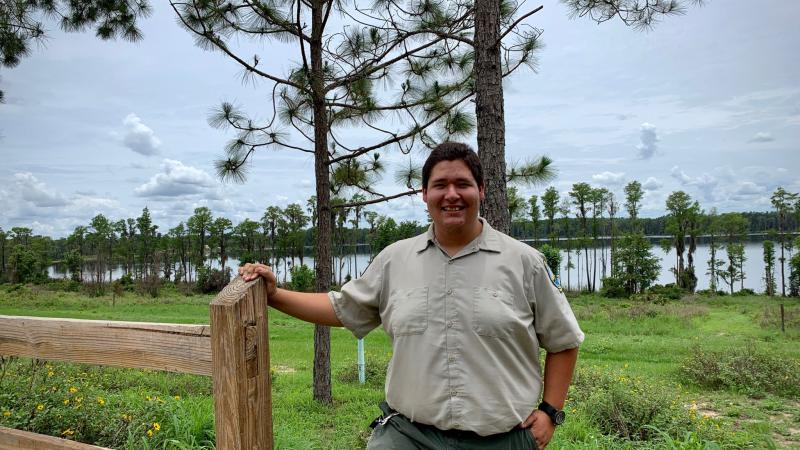 Park Ranger Sam Louzan smiling for the camera with pine trees and a lake behind him