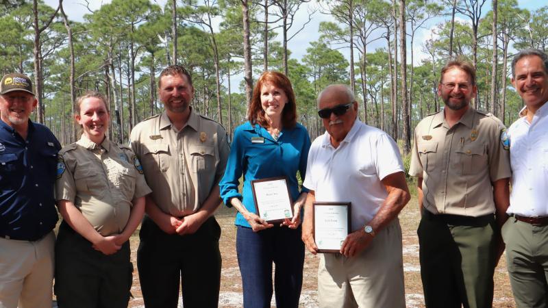 A group of people standing shoulder to shoulder. Two people in the middle are holding plaques. 