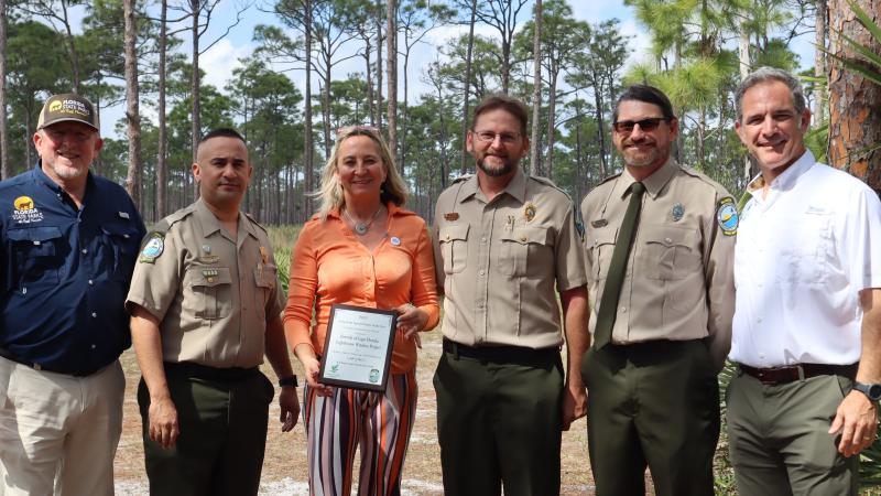 A group of people standing shoulder to shoulder. A woman in the group is holding a plaque. 