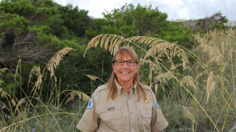 Lynda, smiling at the camera, standing in a field of greenery.