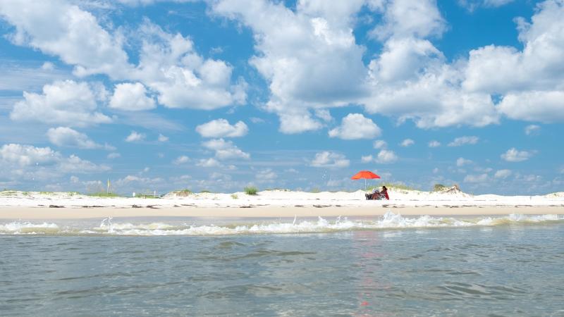 A person sits in a chair with an orange umbrella on the beach at St. George Island State Park.