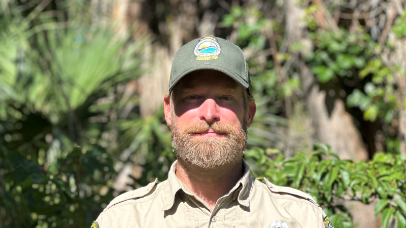 A bearded park ranger standing in front of greenery. 