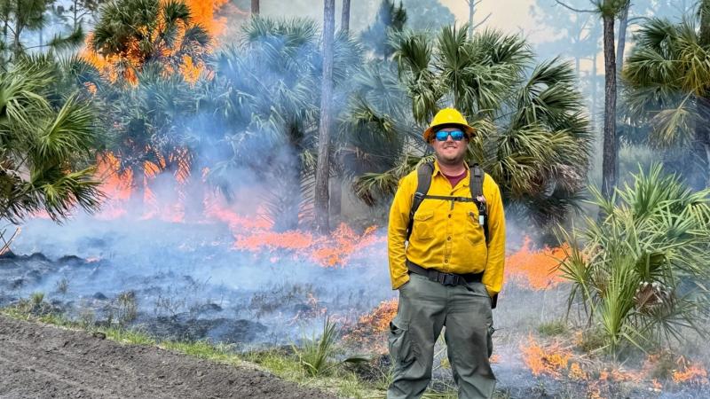 A smiling man wearing protective equipment standing in front of a large prescribed burn. 