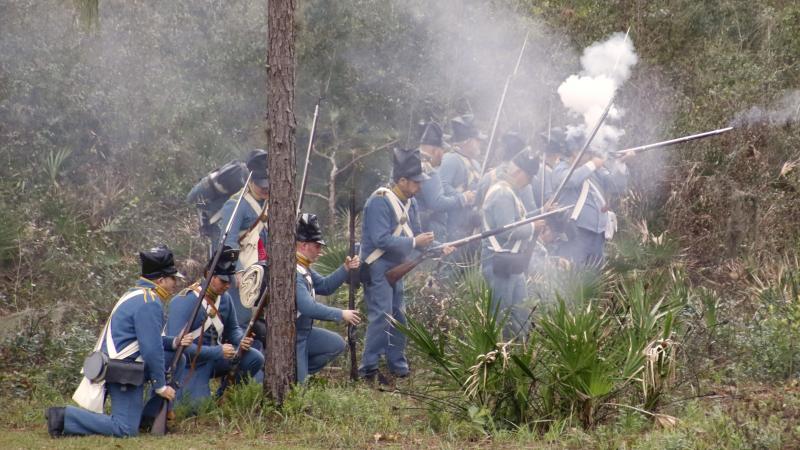 Gun smoke hangs in the air as blue-clad reenactors fire their muskets while crouched in the palmettos.
