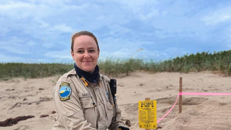 A smiling park ranger writing on a clipboard next to a sea turtle nest.