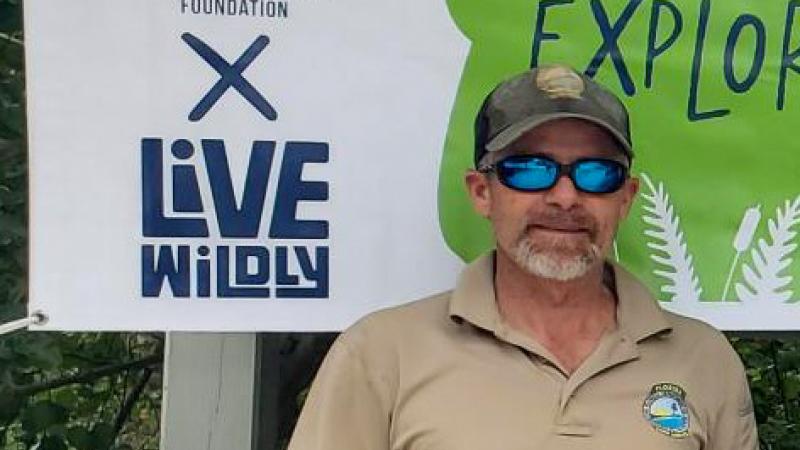 A man with a beard, hat and sunglasses standing in front of a promotional poster. 