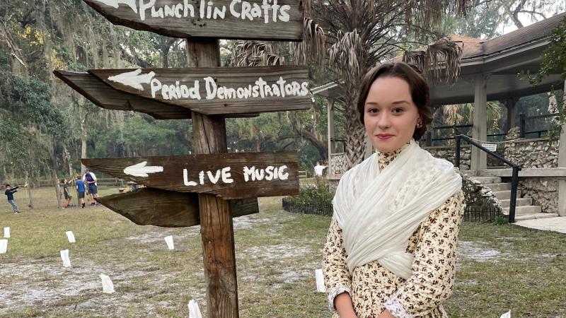 A volunteer in pioneer garb stands by a wooden directional sign at a park event.