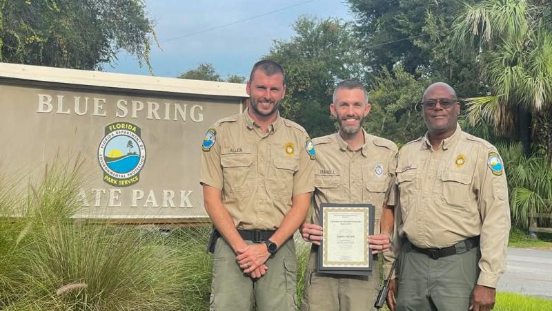 Three park rangers standing in front of a sign that reads "Blue Spring State Park". The park ranger in the middle is holding a framed certificate.  