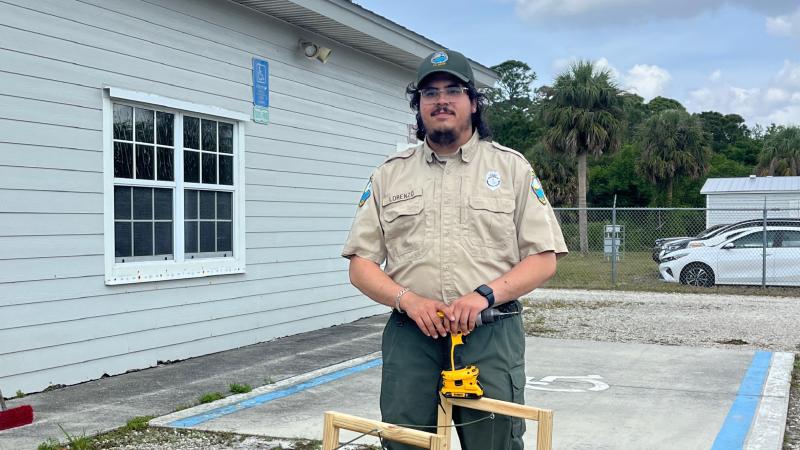 A park ranger holding a drill, standing over a wooden project. 