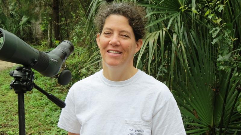 a woman in a volunteer shirt stands next to a spotting scope smiling