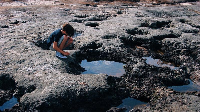 a boy crouches on the edge of a pool formed by hardened black dirt on a beach