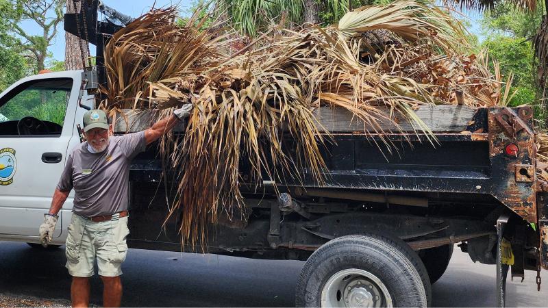 A man standing in front of a pickup with his hand on a pile of palm leaves that is taking up the bed of the pickup. 