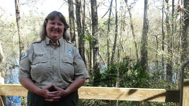 a woman in a park service uniform stands smiling in front of a wet forest.