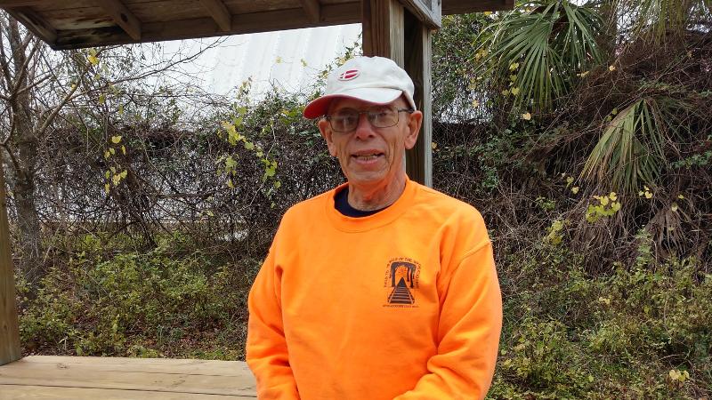 A man in an orange shirt and glasses smiles beside a bench.