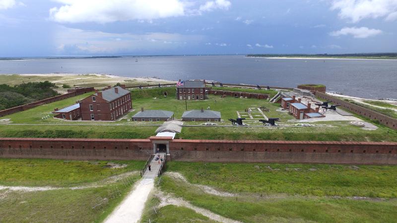 a fort with pentagonal brick walls and numerous building and guns against the backdrop of the ocean.
