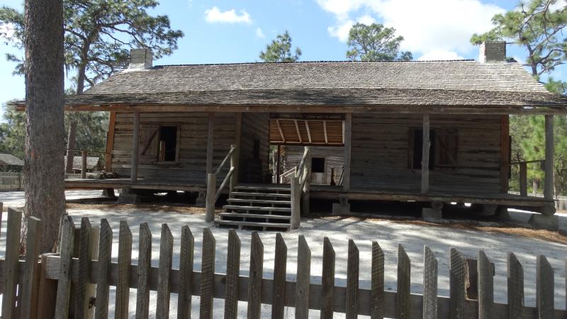 a log cabin with a wood picket fence sit under a blue sky surrounded by pine trees