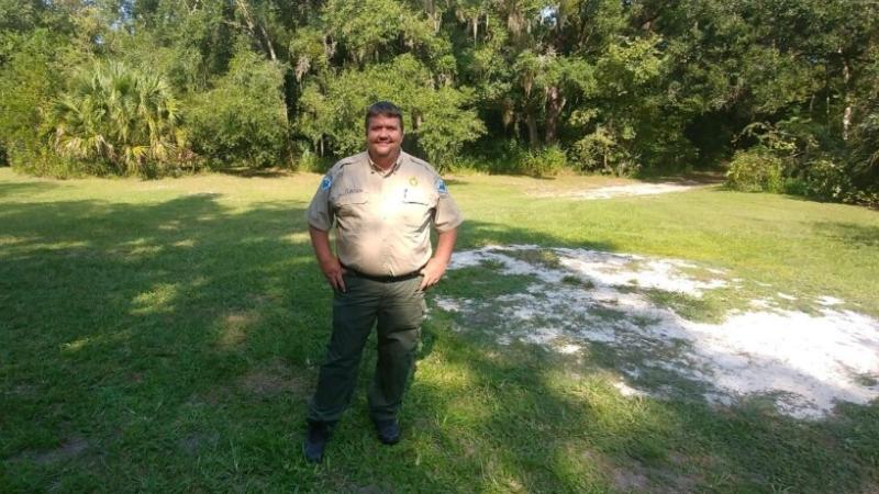 A man in uniform stands in front of a row of trees. 