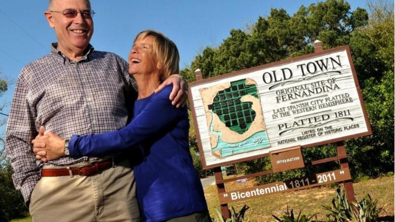 a man and woman hug each other and smile next to a sign for "Old Town Fernandina's Bicentennial"