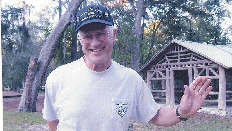 a man in a volunteer shirt and hat smiles and waves in front of a log pavilion