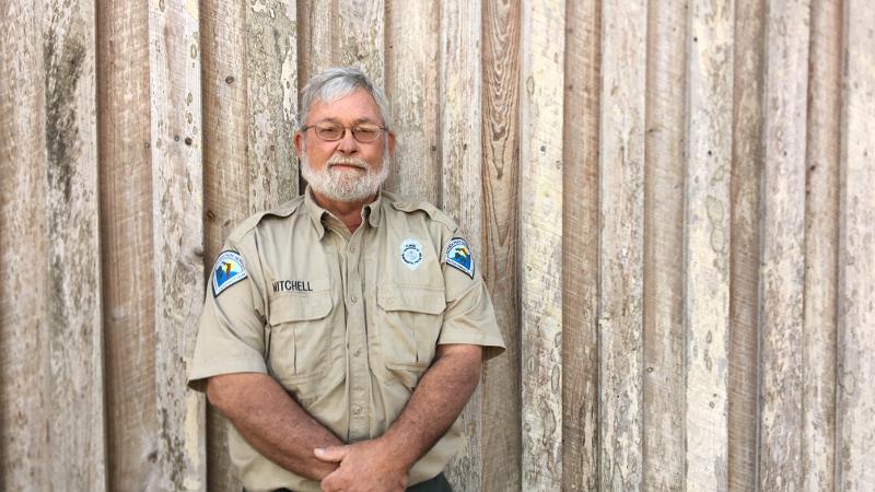 a man in a park service uniform stands against a wooden wall.