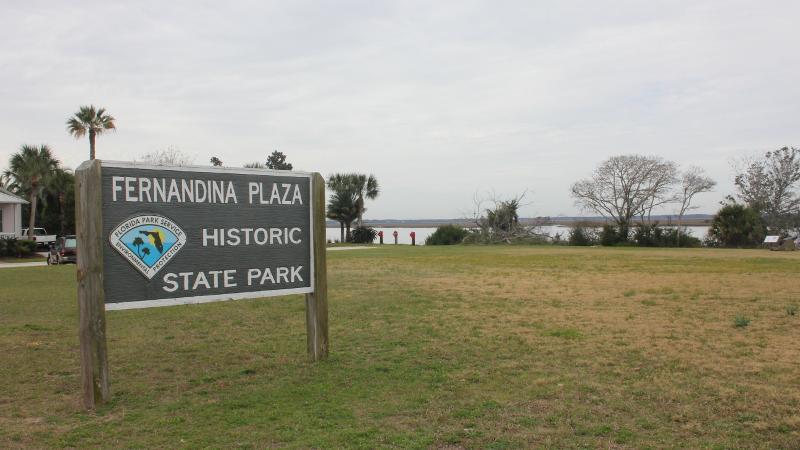 a sign reading fernandina plaza historic state park stands in a grassy field by a river