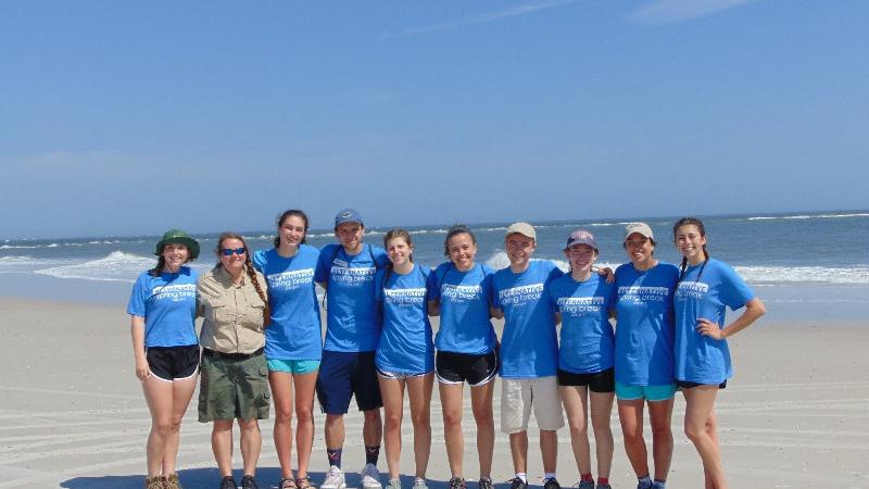 a group of college students in blue shirts and a park ranger stand arm in arm next to each other on the beach
