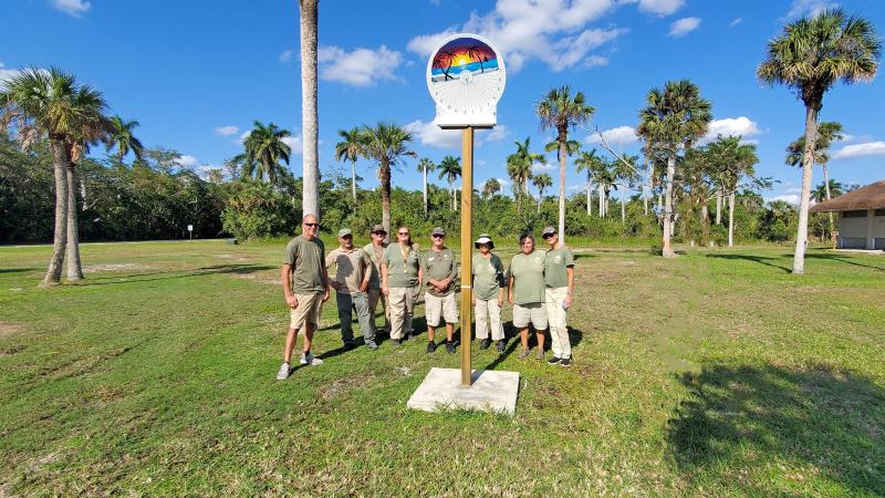 Park volunteers standing next to a sundial with palms, green grass and blue sky