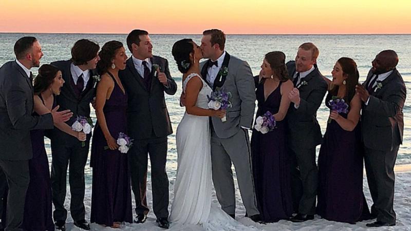 Wedding party surround bride and groom on white sandy beach with glowing orange sunset in the background.