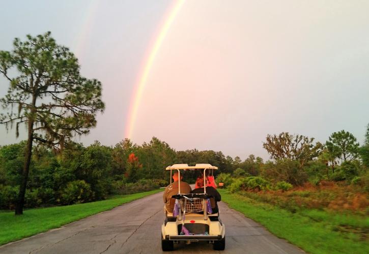 A view of people riding in a golf cart with a rainbow in the background across the sky.