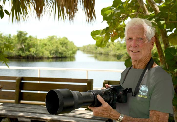 man holding a camera near a river and park bench