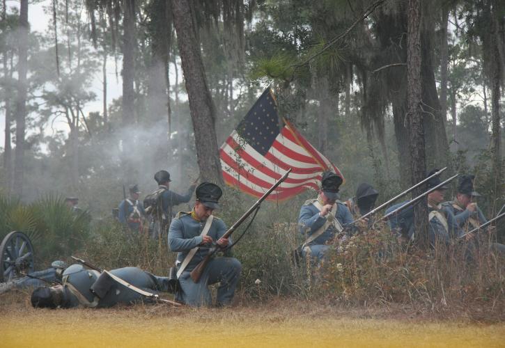 A group of reenactors in uniform, shooting their muskets, with the flag flying through the smoke.