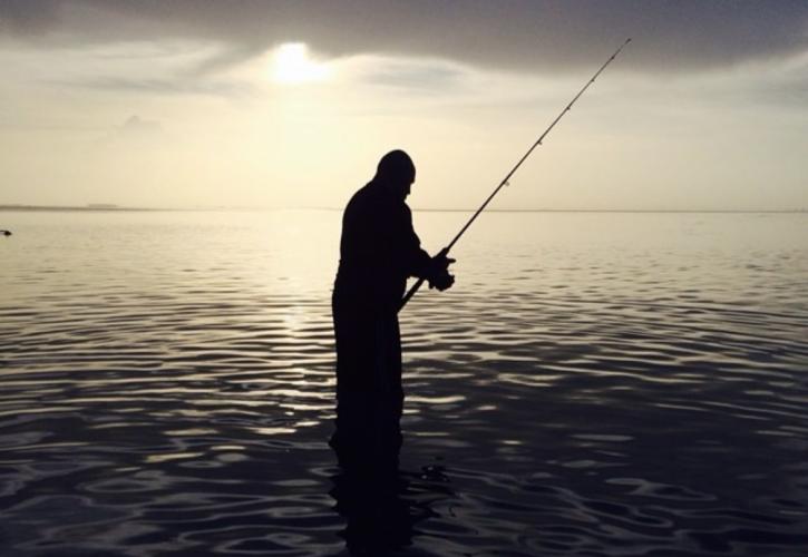 A silhouetted person casting into a calm, grey ocean.