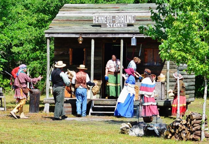 A view of a group of reenactors dressed in period clothing in front of a wooden building.