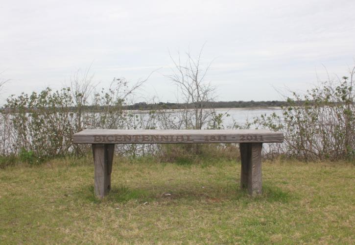 a wooden bench with "Bicentennial 1811-2011" carved in the seat sits in front of a river.