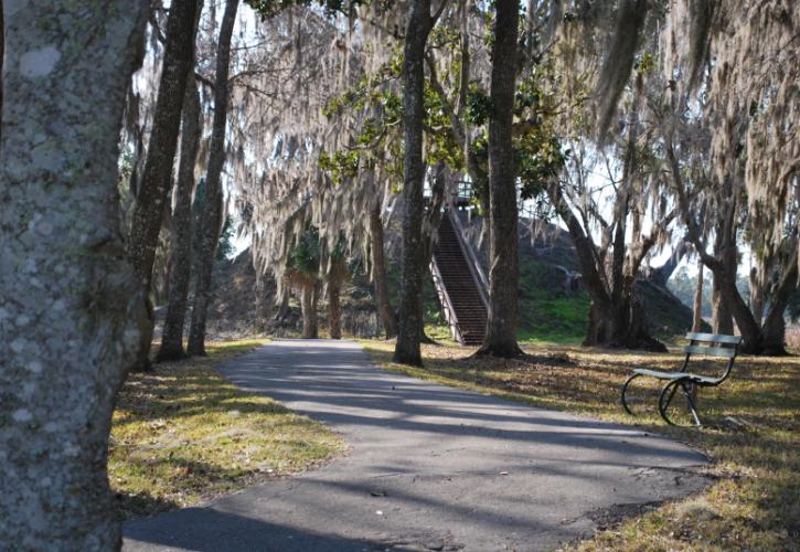 a paved path winds towards a large hill with stairs. A bench and many trees line the path.