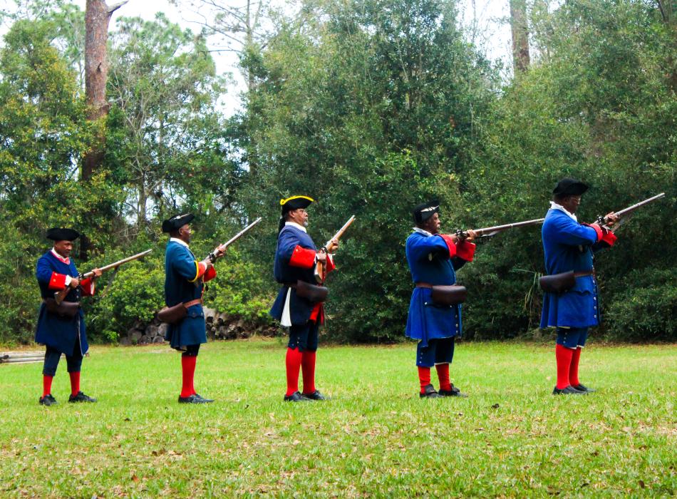 A line of 5 reenactors wearing blue and red historic costume prepare to fire muskets.