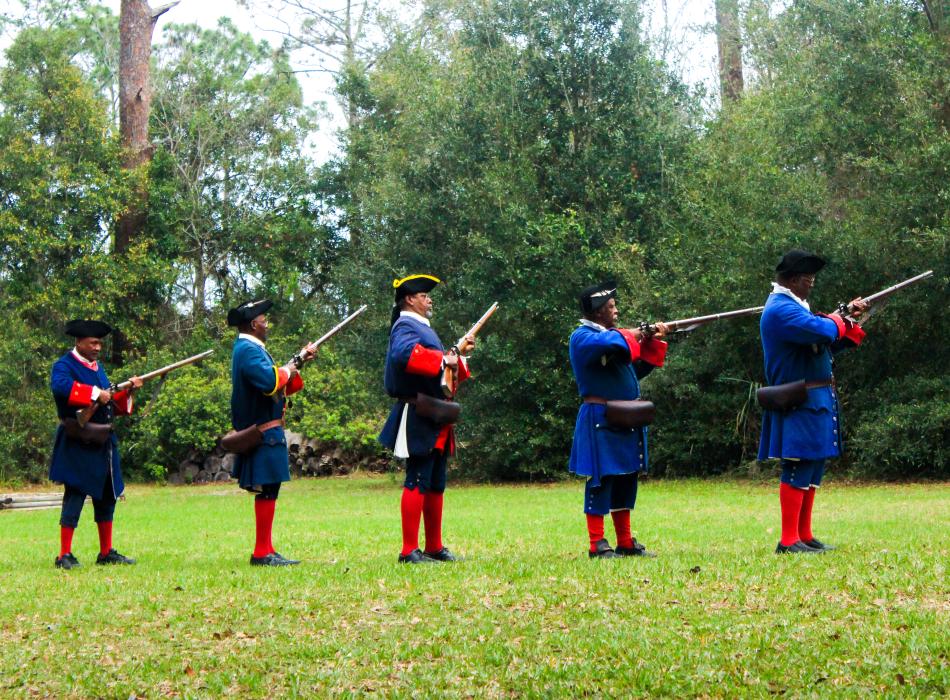 A line of 5 reenactors wearing blue and red historic costume prepare to fire muskets. 