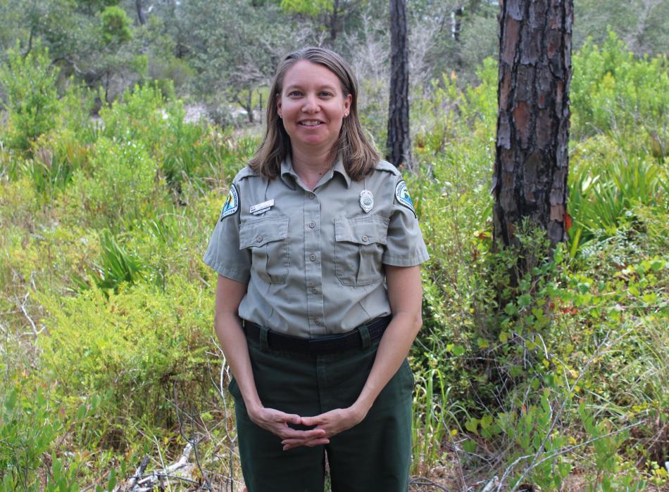 Park Ranger stands in middle of lush green vegetation and pine trees. 
