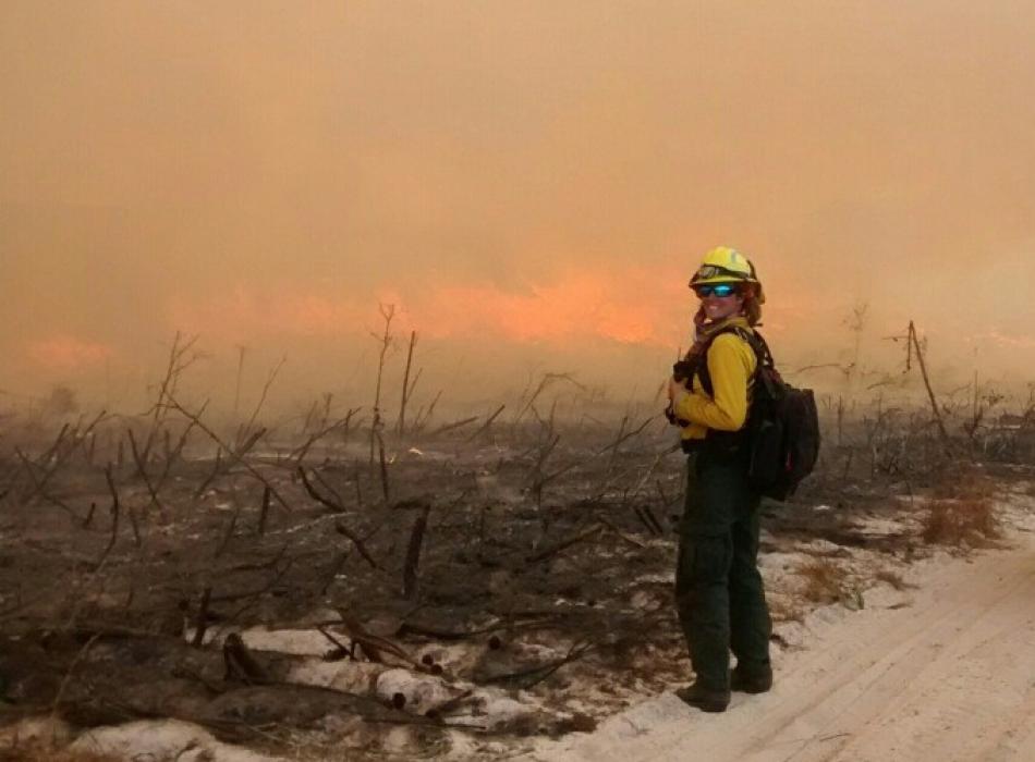 A lady with fire gear on stands infront of land that has just been burned.