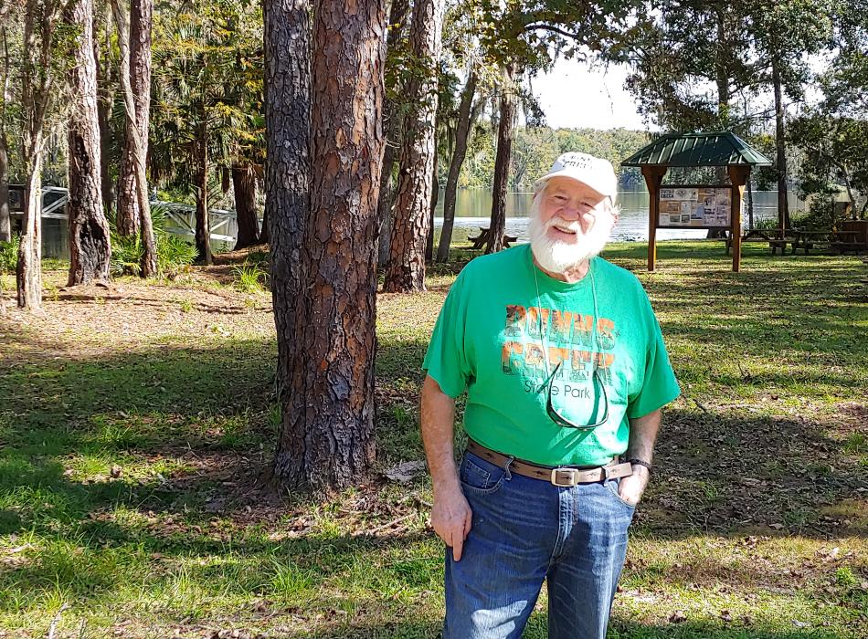 A man with a green shirt and a tan hat stands in front of trees.