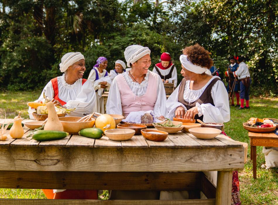 Three Black women in historic costumes talking over a table of food.